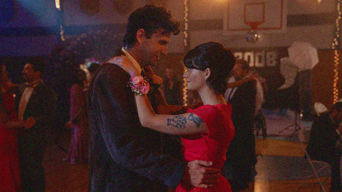 A thirty-something man and woman dance center frame at a prom held in a high school gym.