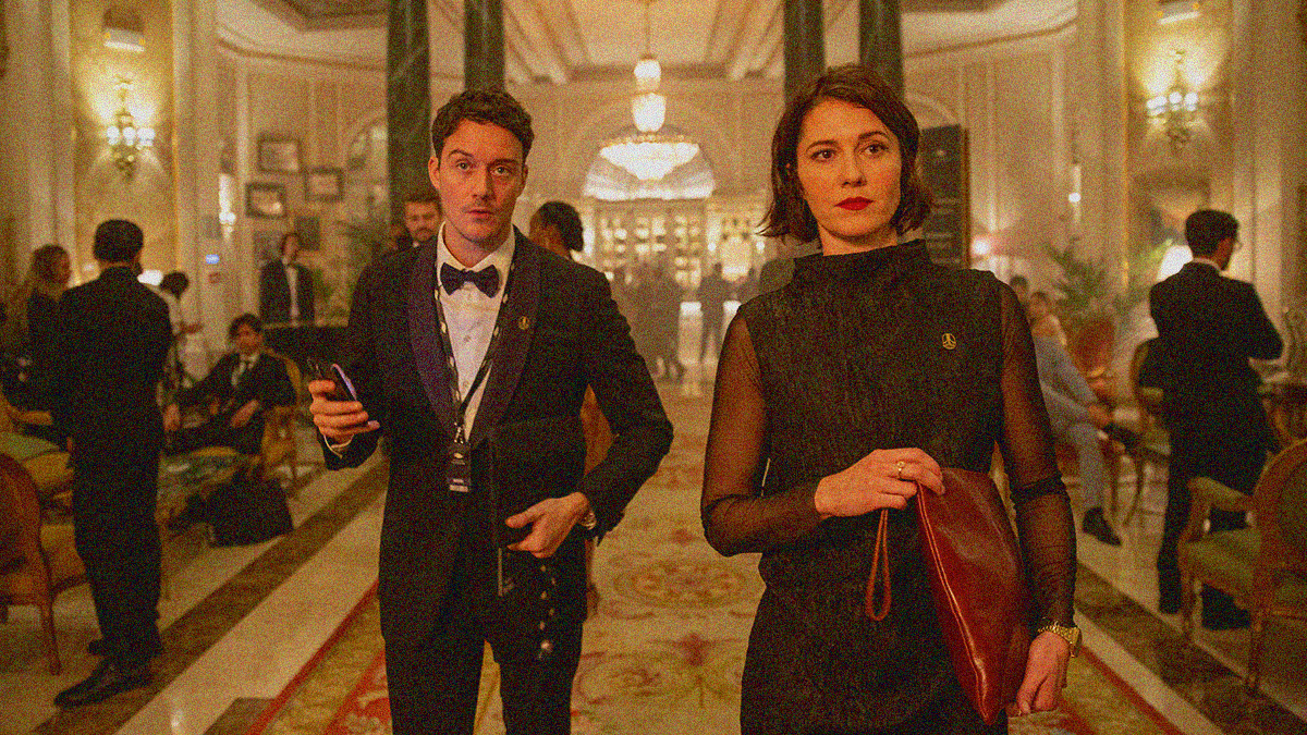 A woman (right, foreground) and a man (left, a little behind her) walk towards the camera through an ornate lobby with other people in formal wear.