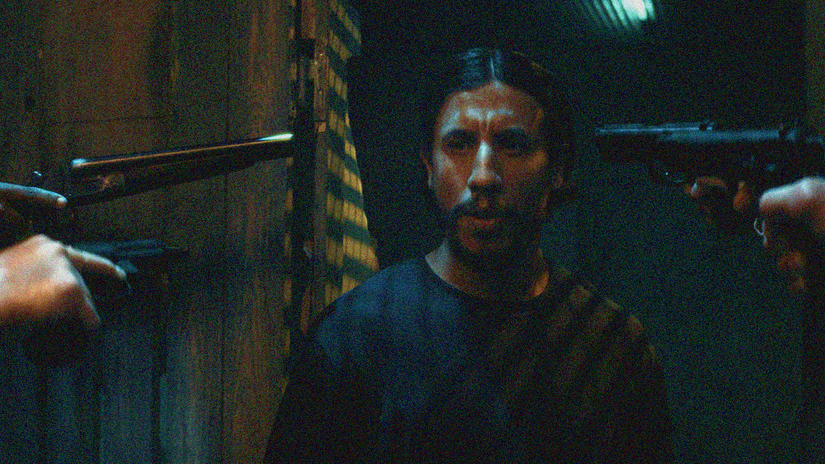 A man at center of the frame looks off-camera while three guns (two from left and one from right) enter the frame pointed at his head.