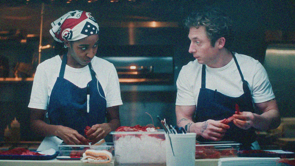 A woman with wrapped hair and a man with tattoos stand side by side at a kitchen counter with prawns, looking at each other with intent.
