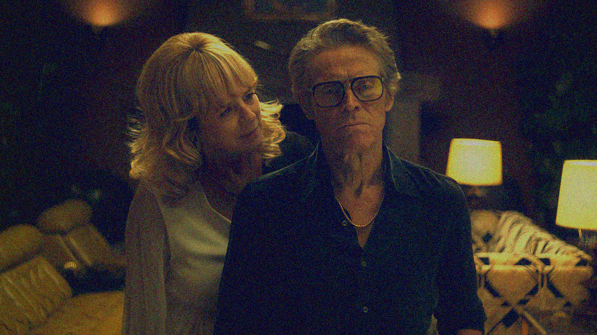 A man in a black shirt, glasses, and gold chain stares past the camera with a stern look. A woman stands just behind him, head tilted to look his way and speak.