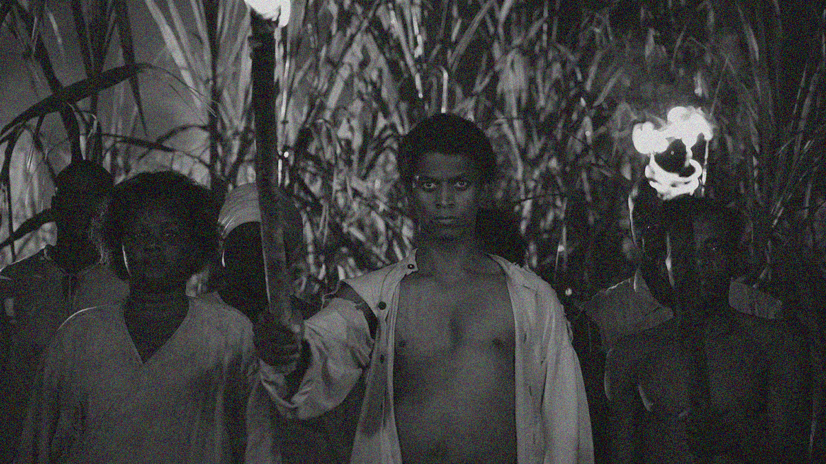 Black and white image of a group of Black men and women standing with lit torches in a sugar cane field.