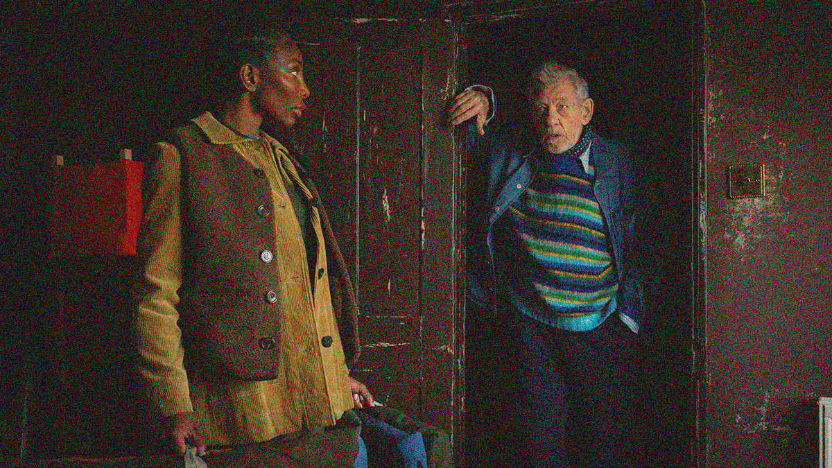 A woman in a vest carrying cloth-wrapped paintings peers at an older man in a sweater leaning in a door frame speaking to her.