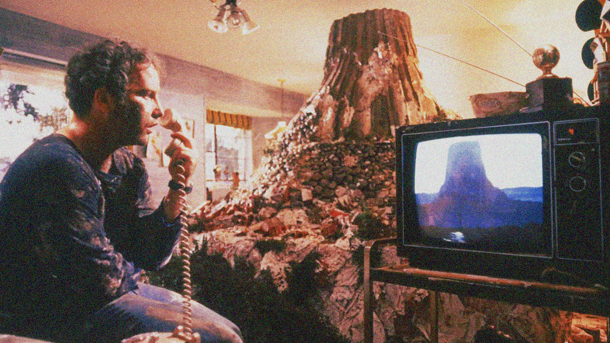 A man looks dumbfounded at the image of Devils Tower National Monument on the television while holding onto a phone receiver. His unwitting model made from dirt and stone of the same image is seen rising in the background from its place in his living room.