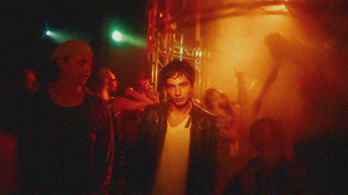 A man staring into the camera seems to almost be floating at the center of a raucous rave seen shrouded in orange.