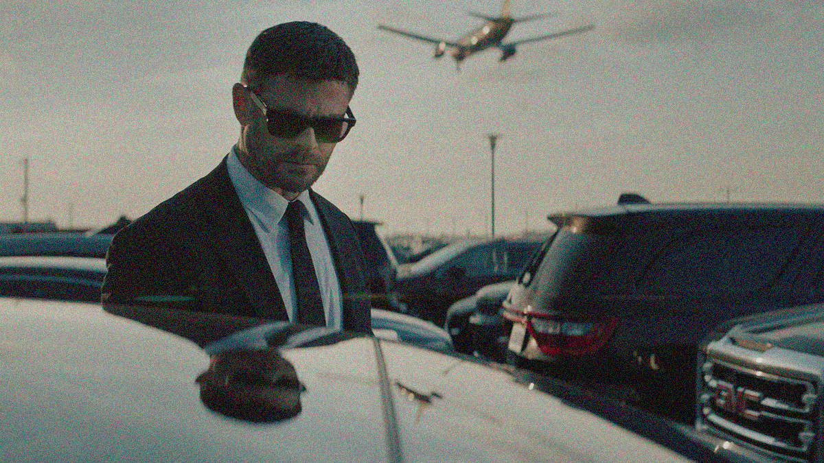 Close-up of a man in suit and sunglasses is seen looming over a car in an airport parking lot. A plane is seen in the sky above him.