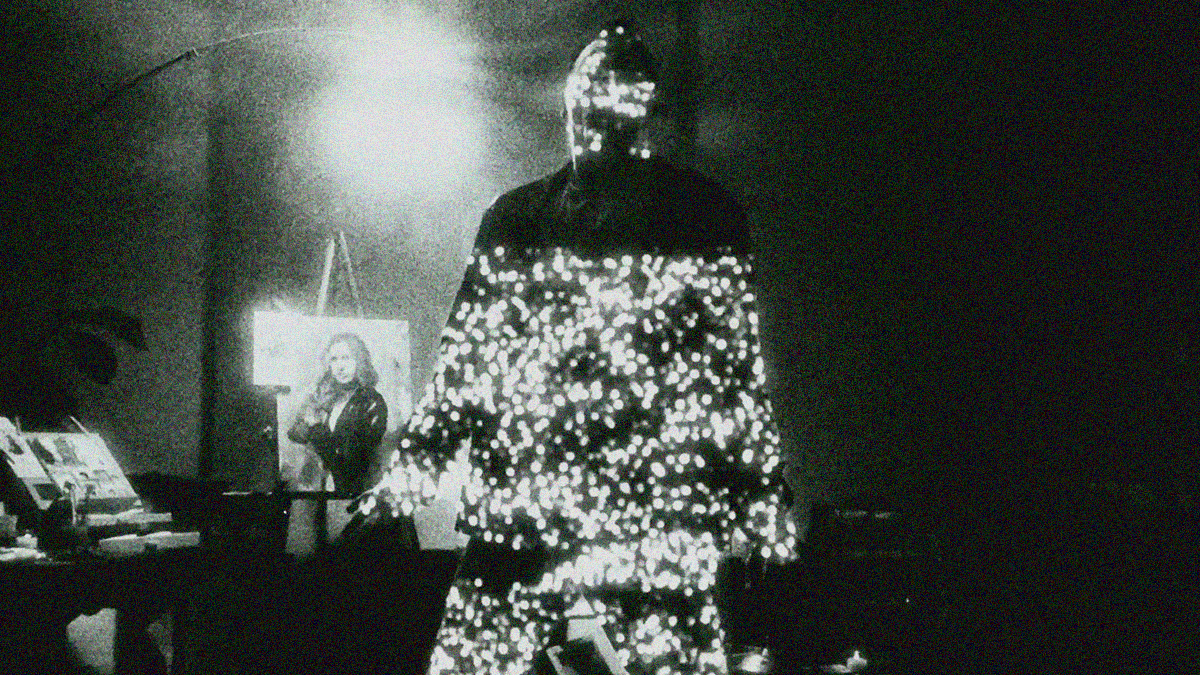 The figure of a man is shrouded in darkness while a field of tiny lights are projected upon him. A painting of a woman on an easel is seen behind him.