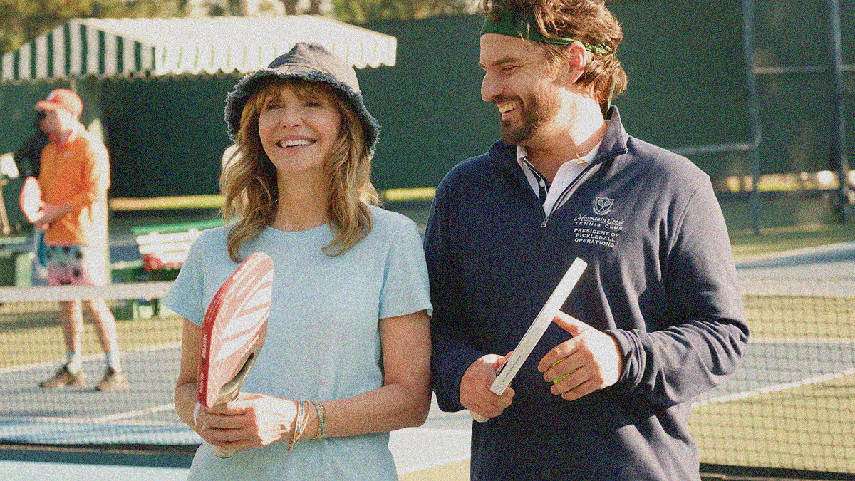 A woman and man stand side-by-side smiling on a tennis court with pickleball rackets in-hand.