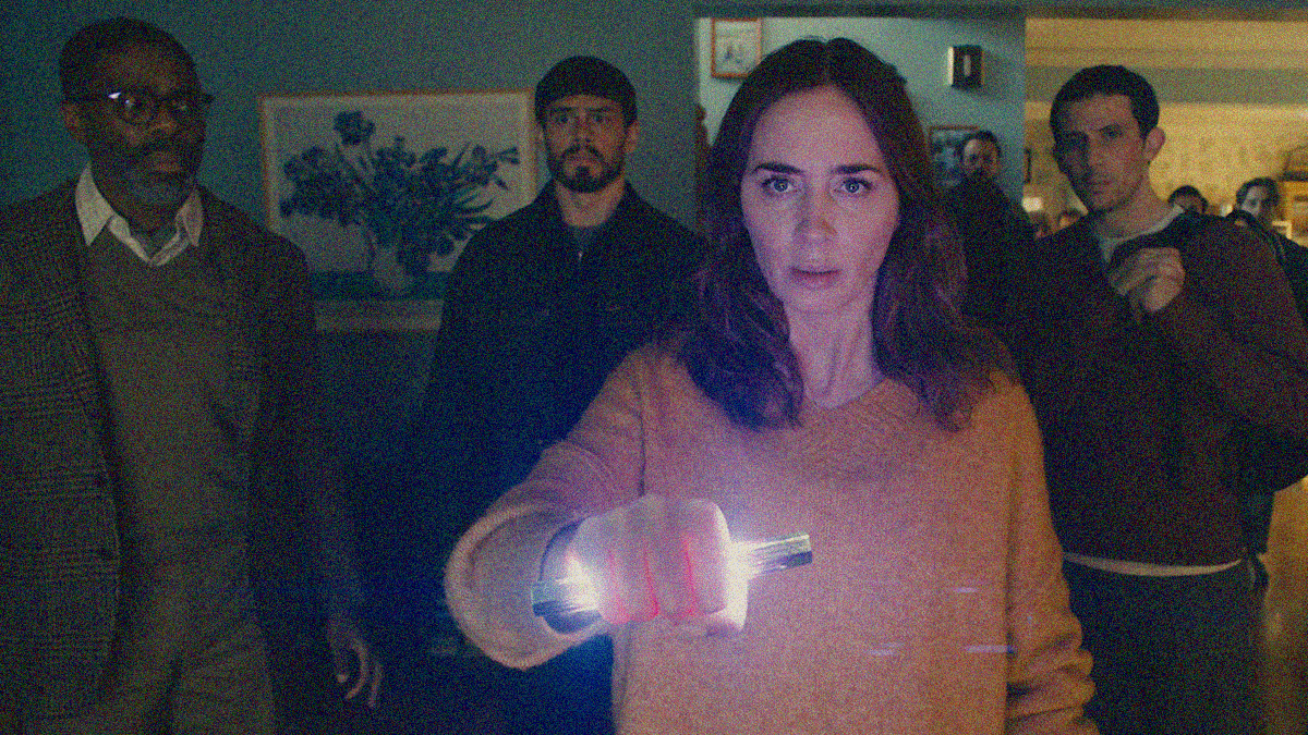 A woman in the foreground stares ahead while holding a glowing object. Three men stand right behind her watching with more behind them.