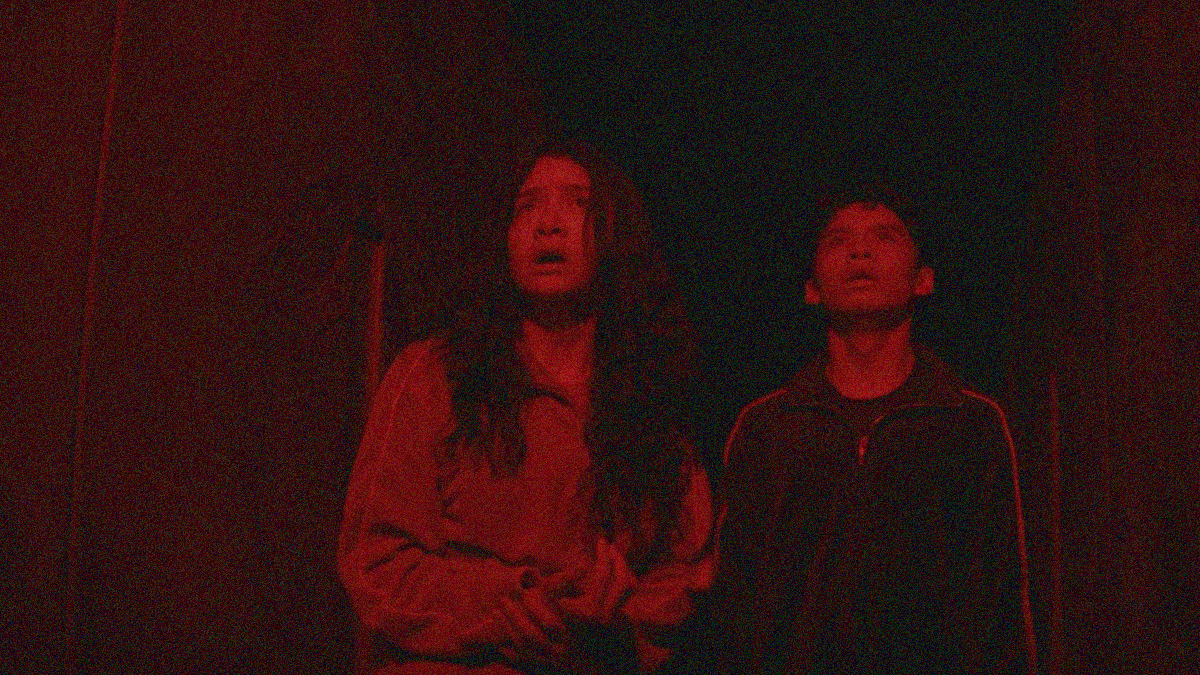 Red-tinted image in darkness of a girl and boy looking upwards with dismay.
