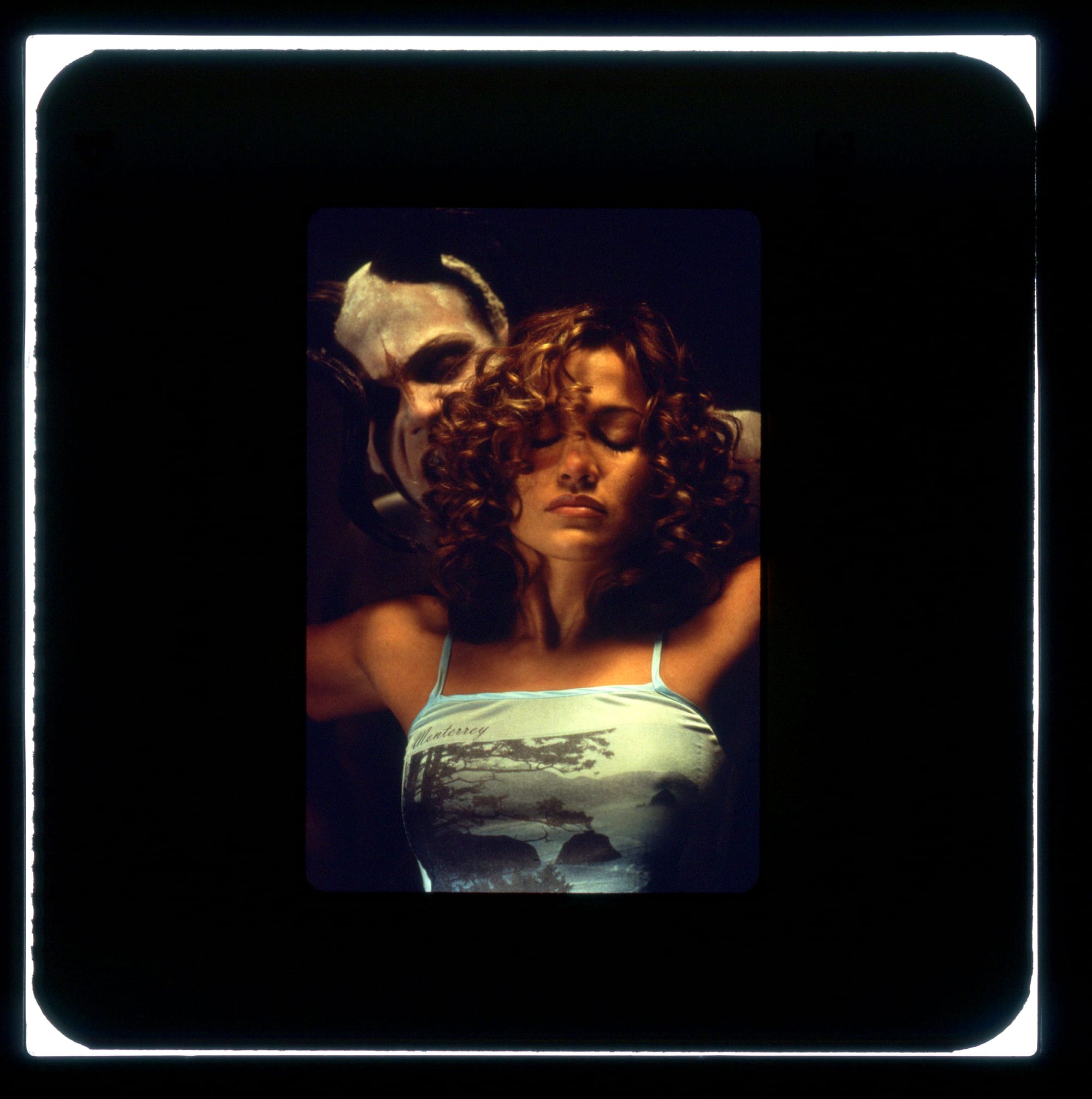 Close-up of a woman with curly hair from chest up. Her eyes are closed and her arms extend off-screen to bend behind her towards a sinister man with white-painted face and horns.