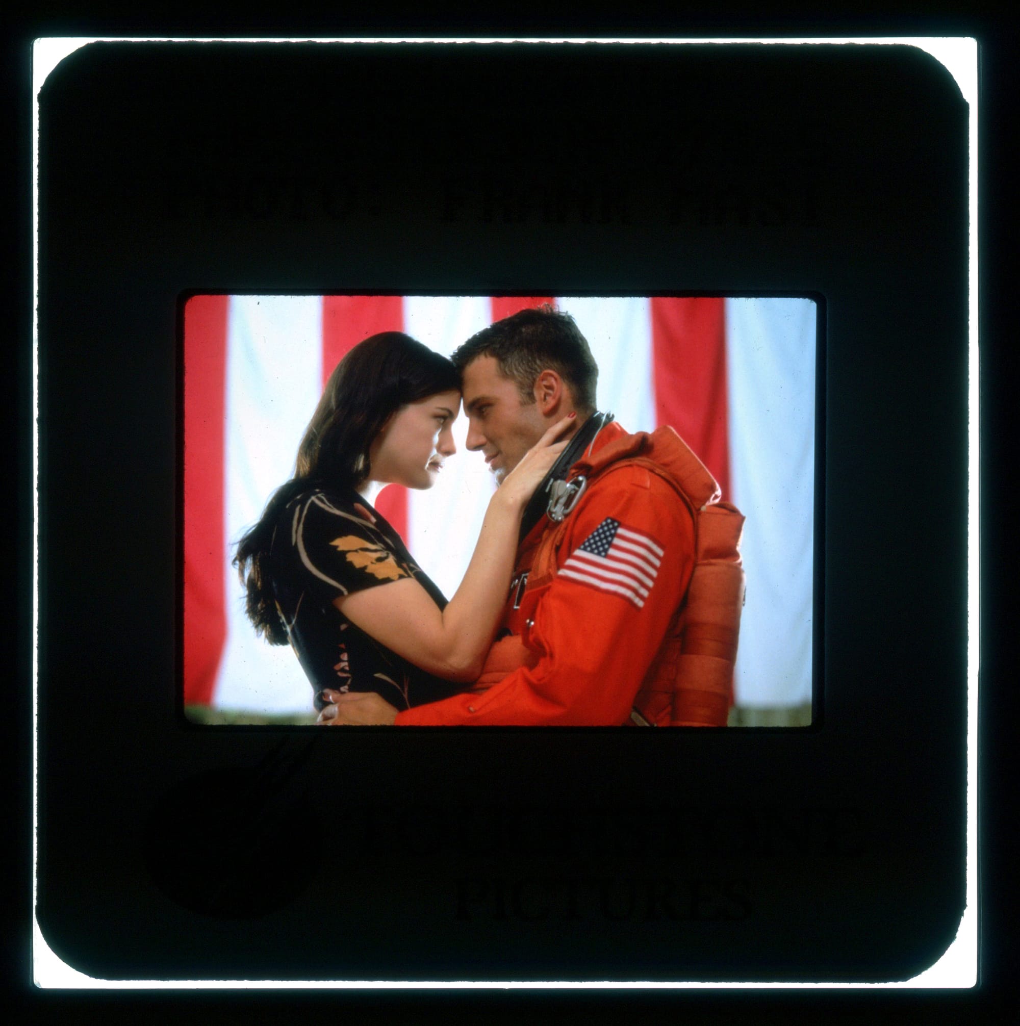 Color publicity slide. Profile shot of a woman and man facing each other. She's wearing a black dress with floral pattern, he is wearing an astronaut uniform. His hands are on her waist, her hands are on his neck, their foreheads are touching as they look in each other's eyes.