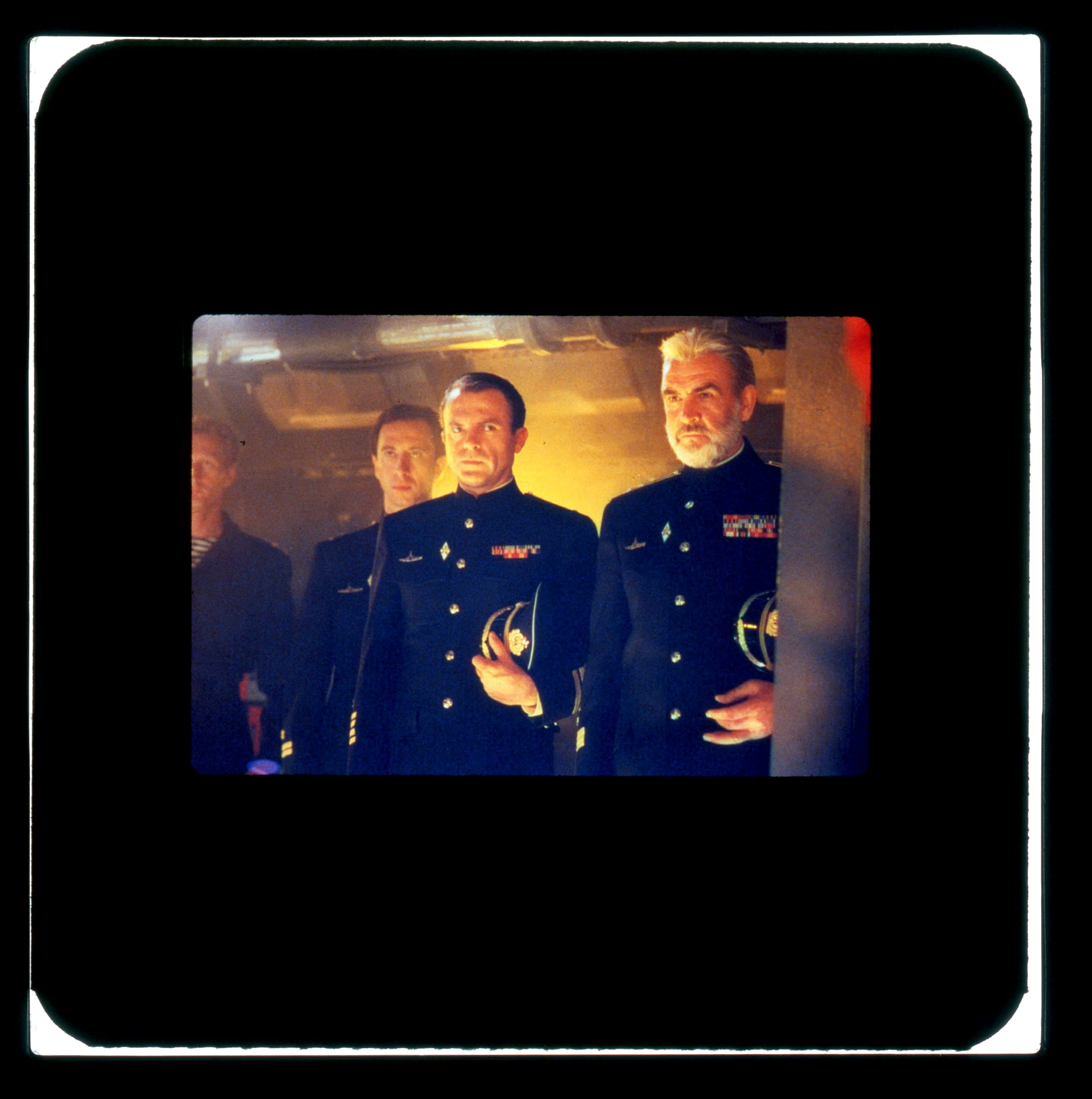 Three men in uniform are seen facing left with caps under arms, but the man in the middle is looking right.