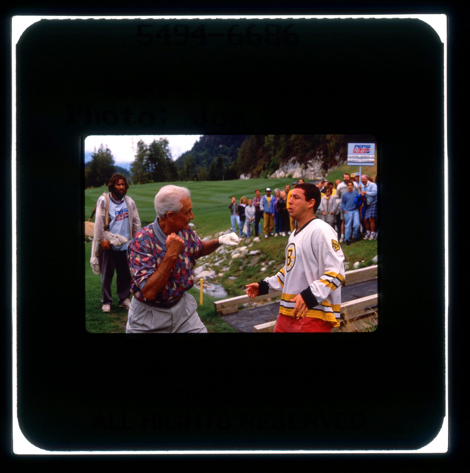 Color publicity slide of Bob Barker getting ready to punch Adam Sandler on the golf course. Allen Covert is in background on left holding Sandler's clubs. A crowd is watching in the background at right.