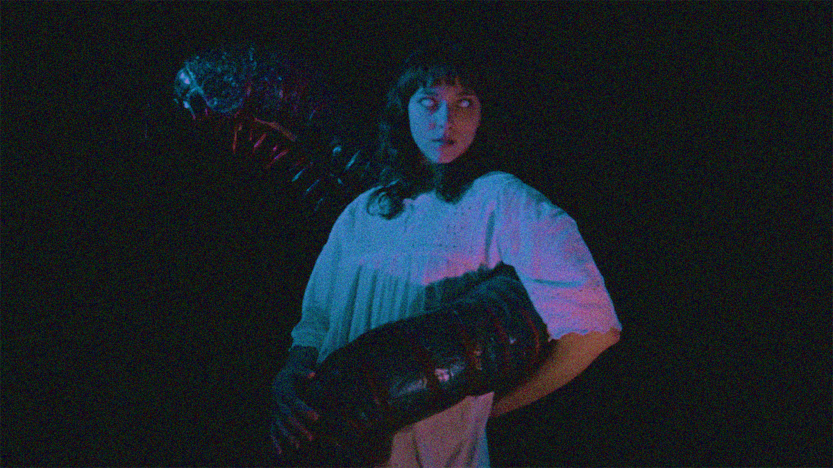 A woman in a night gown with white eyes stands center frame with a giant insect wrapped around her.