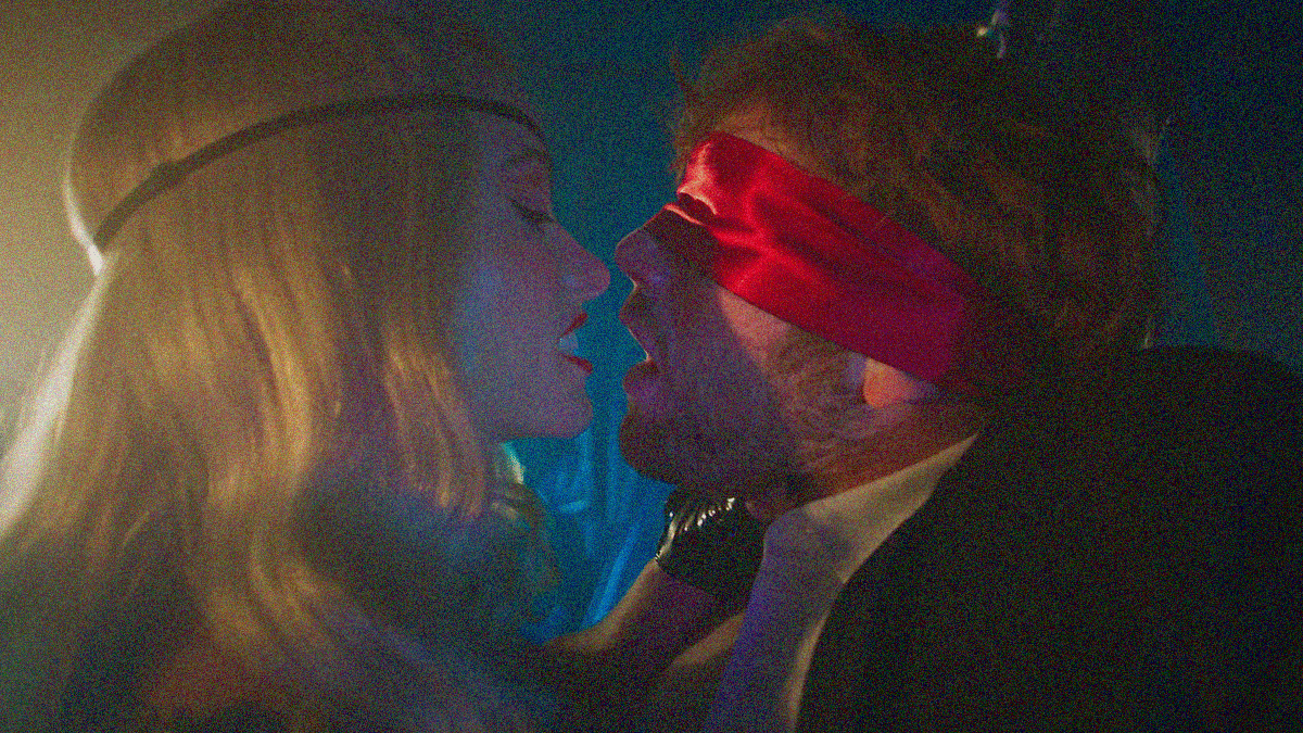 Close-up of a blonde women baring teeth as she goes in for a kiss with a man in a red blindfold.