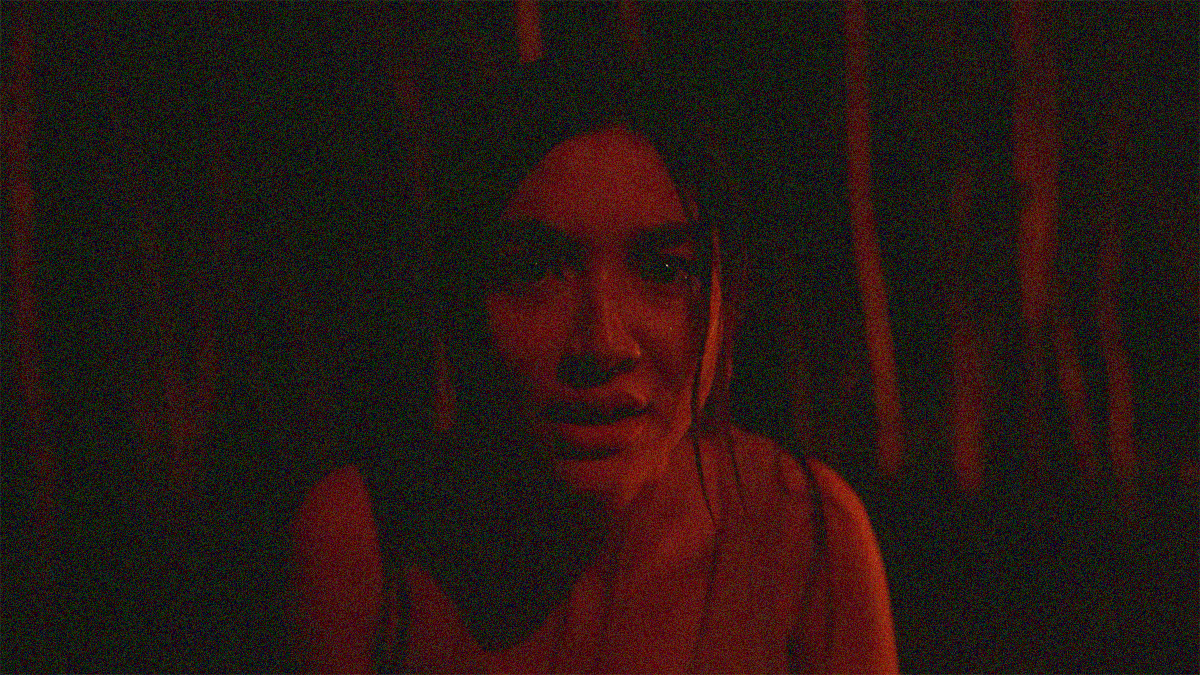 Close-up of a woman in a dark forest tinted red. She's looking directly into the camera.