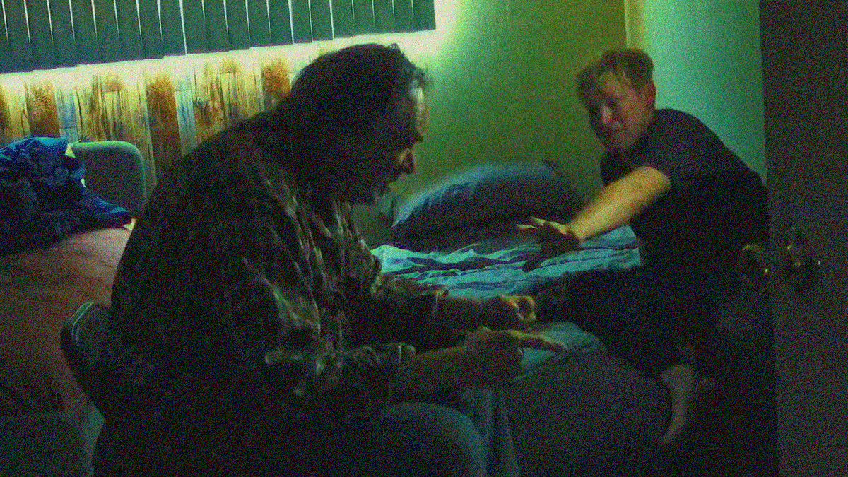 An older man sits on a chair in angry distress while a younger one looks scared while reaching out to stop him from doing something.