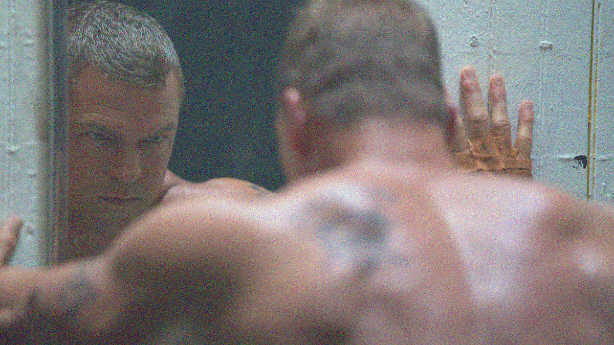 Close-up from behind of an imposing, shirtless man looking at himself in a mirror while pressing his hands on either side of it.