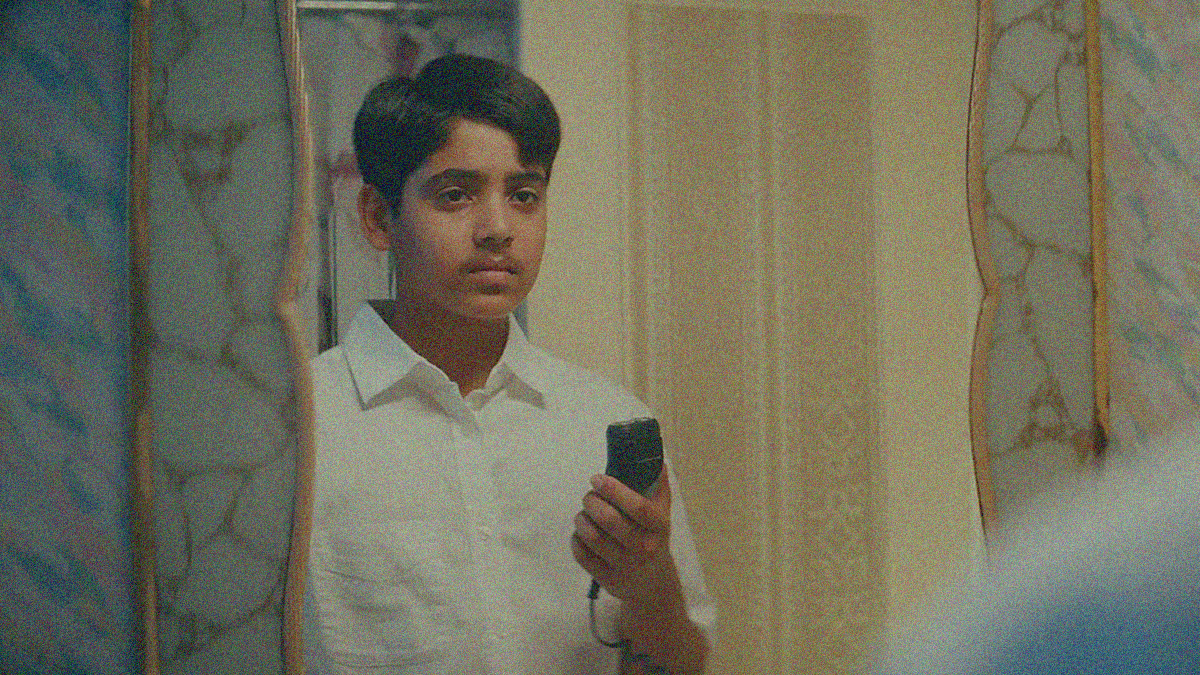A teen boy with a mustache looks into a mirror while holding an electric razor.