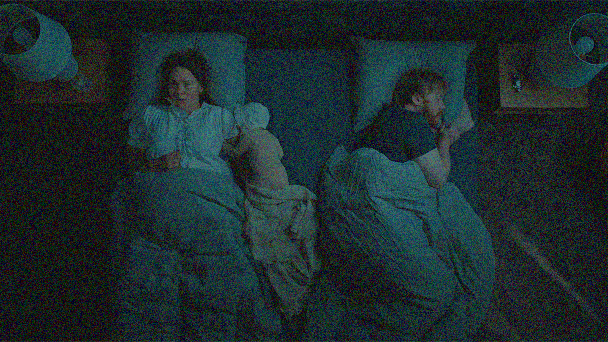 Overhead shot of a darkened bedroom. A woman on left lies on her back awake with a baby curled at her arm. A man at left sleeps on his side.