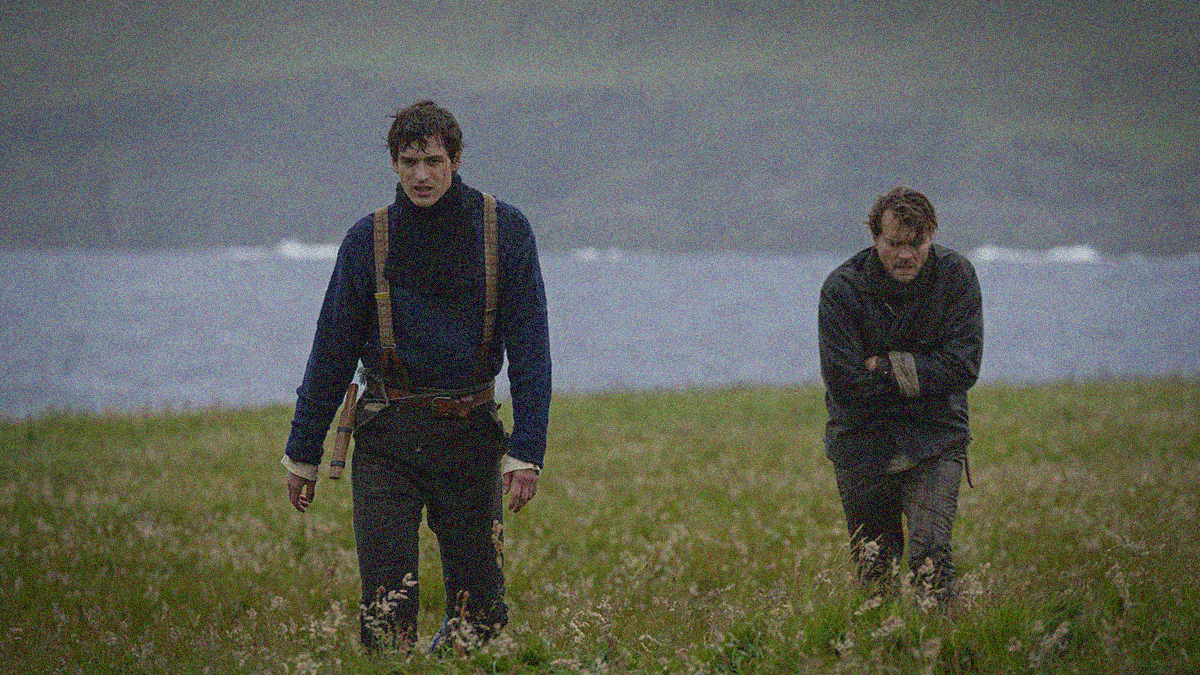 Two men against a scenic backdrop of wall and rock cliffs. Man on left stands straight walking towards camera. Man on right is hunched over, arms folded.