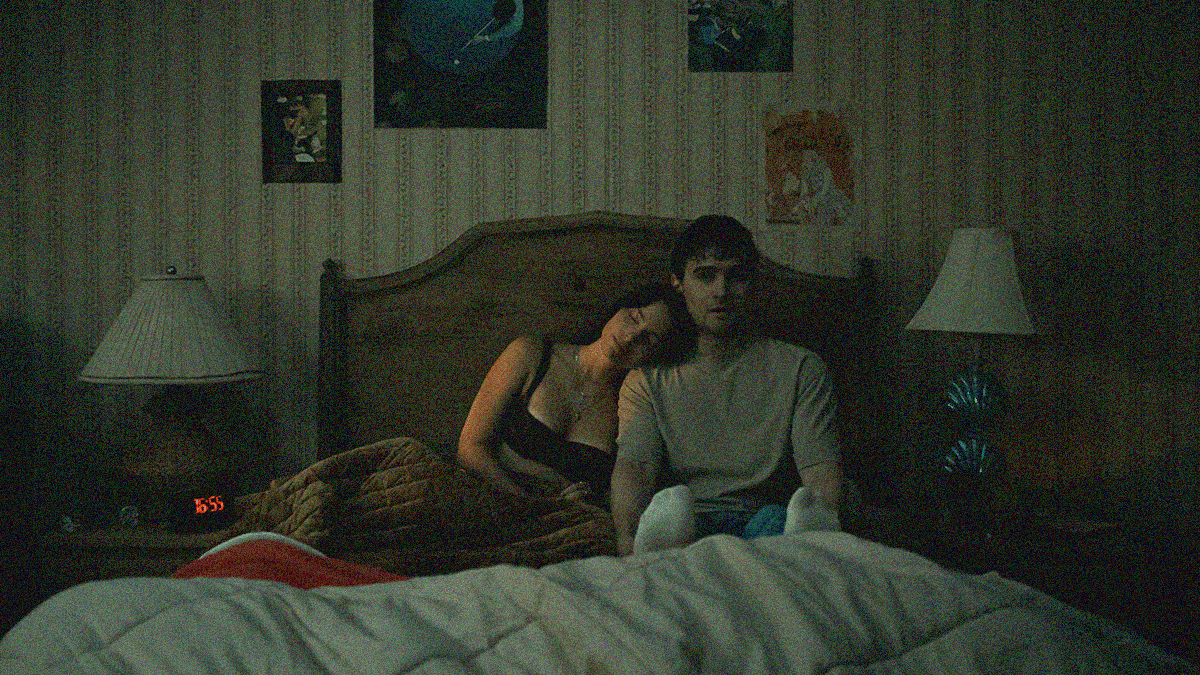 A woman in a bra with legs under the bed covers rests her head on the shoulder of a man sitting on top of the covers and looking spooked.