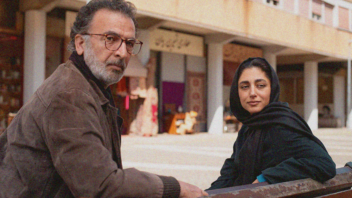 A man with grey beard and glasses (at left) and a woman in hijab (at right) sit on a bench outside. They are both turned around to look off frame towards us over the backside of the bench.