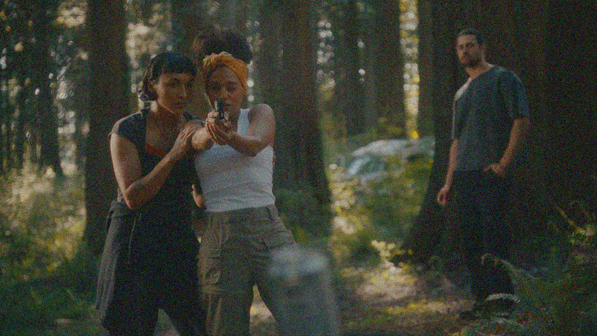 A woman in the midground of a forest scene at left is helping another woman point a gun at the blurry beer can at the foreground. A man stands watching in the background at right.