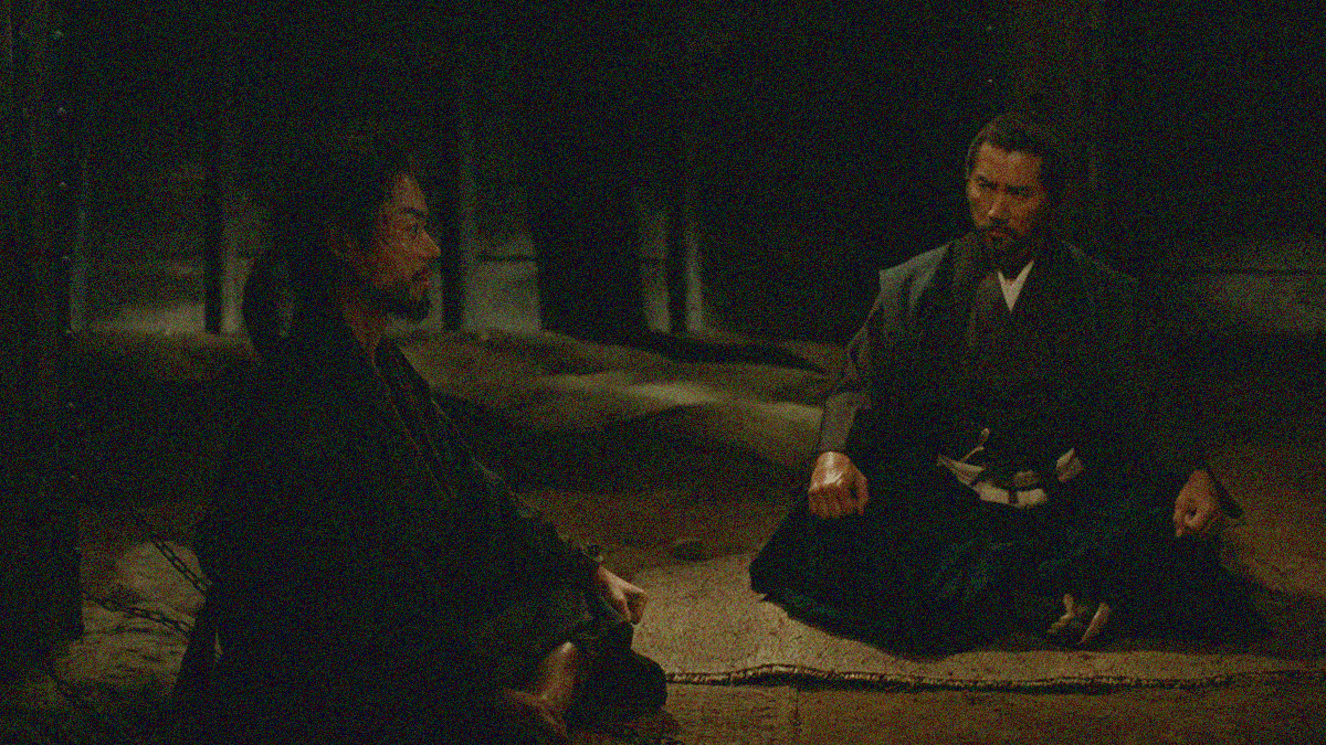 Two men sit crosslegged on the floor of a dungeon. The one on the left is in chains facing right. The one on the right looks at him intently.