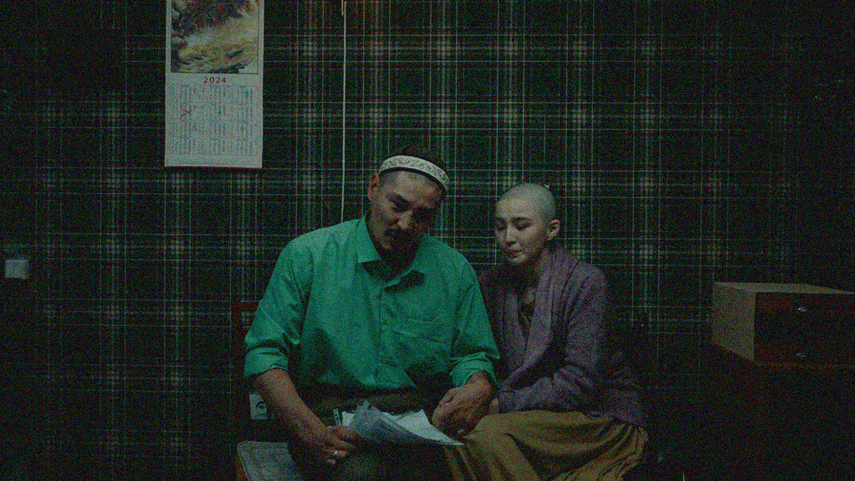 A man wearing a kufi over his shaved head sits besides a woman with a shaved head. Both are looking at the camera in distress. He is holding papers.