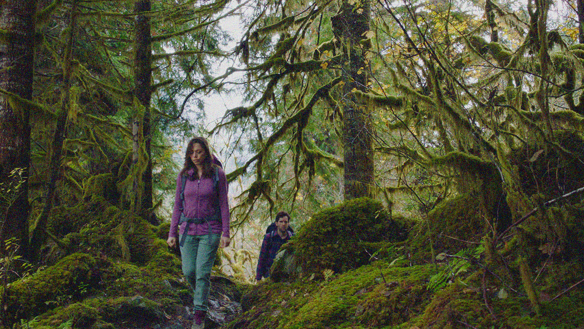 A woman is in the foreground walking between trees in a forest. A man is seen further behind her, just coming into view from behind a rock.