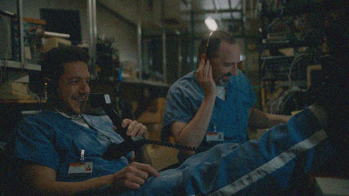 Two men in blue jumpsuits are sitting and laughing at a desk of communication equipment. The one on the left has a mic to his mouth.