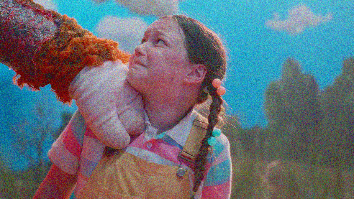 A young girl with pigtails looks scared as a mascot's hand reaches into frame to grab her shoulder.