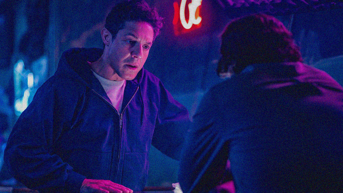 Two men sit inside a blue-tinted bar. One has his back to us. The other looks at him with distress.