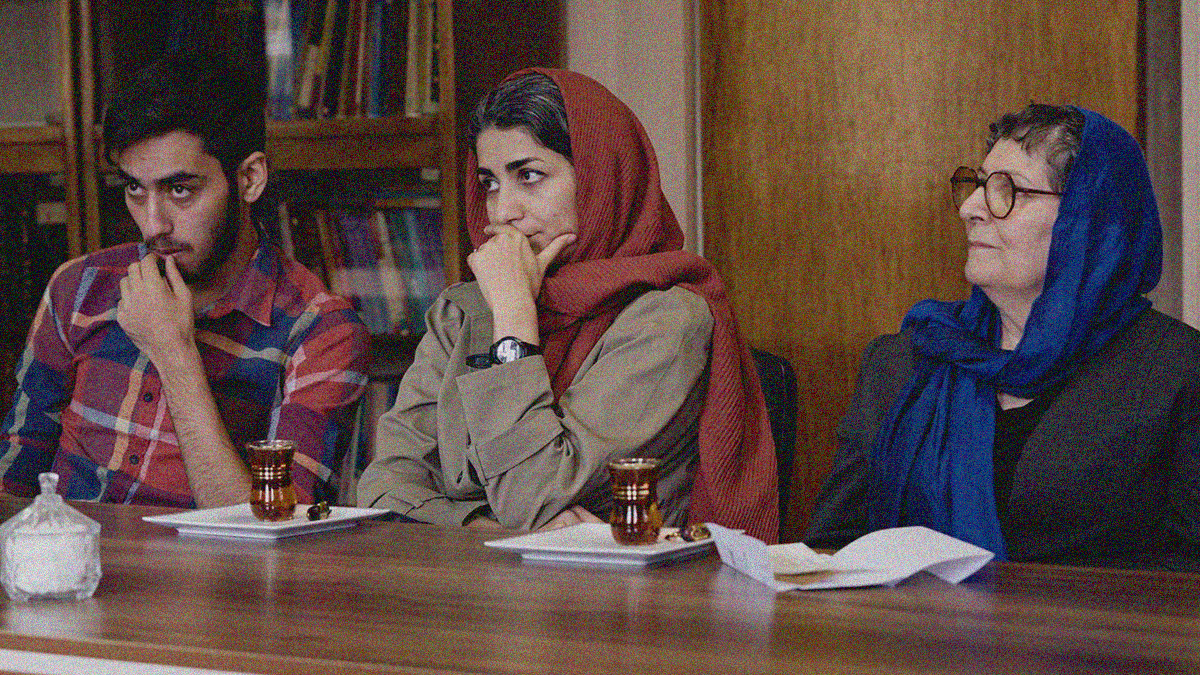 A man and two women sit at a table looking off-screen left while listening to someone out of frame.