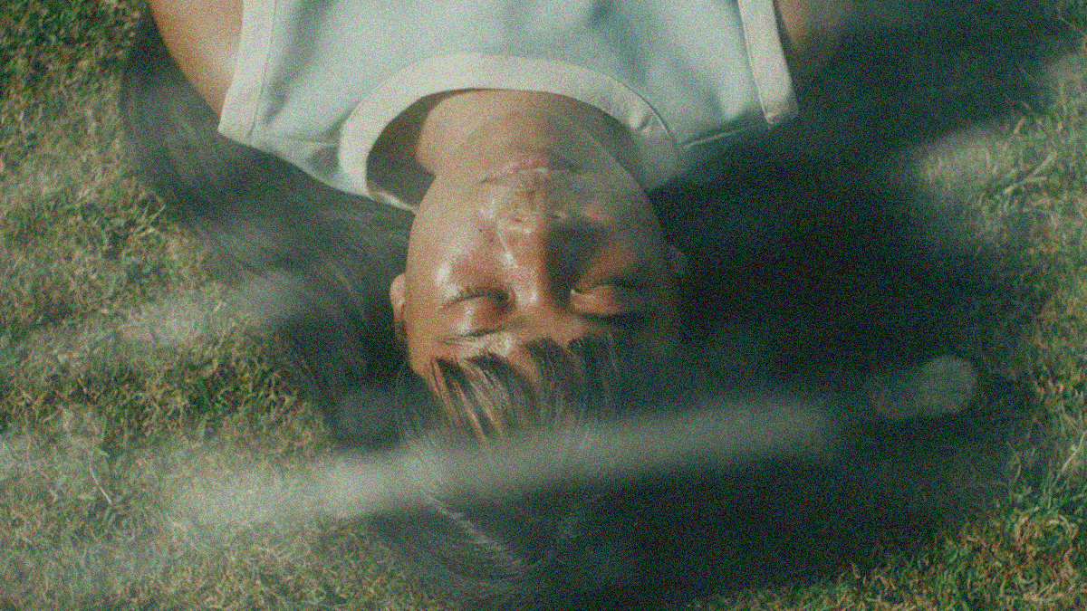 Overhead close-up of a young woman lying on green grass with her eyes closed. It looks like streams of water from a sprinkler are shooting above her face.