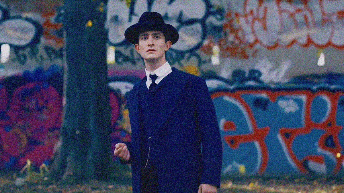 A man in a suit, tie, and hat from the early 1900s looks very out of place at the center of the frame considering a wall of modern graffiti is seen behind him.