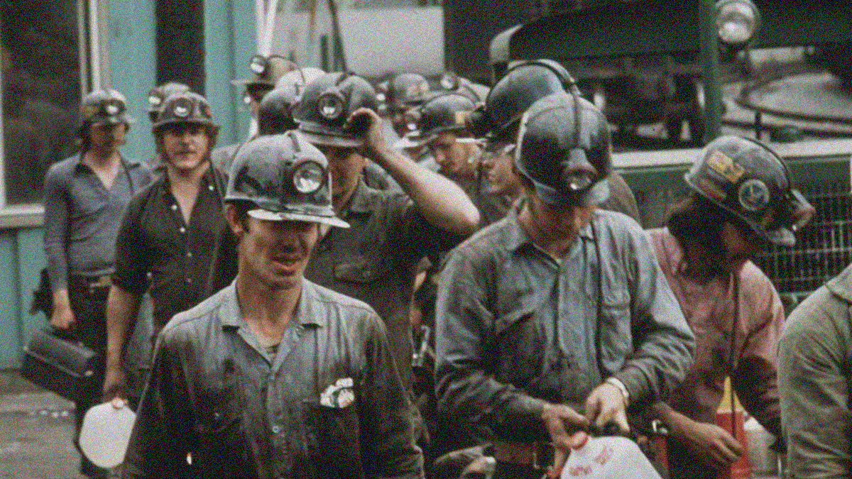 A group of miners in helmets and carrying lunch pails walk towards the camera.