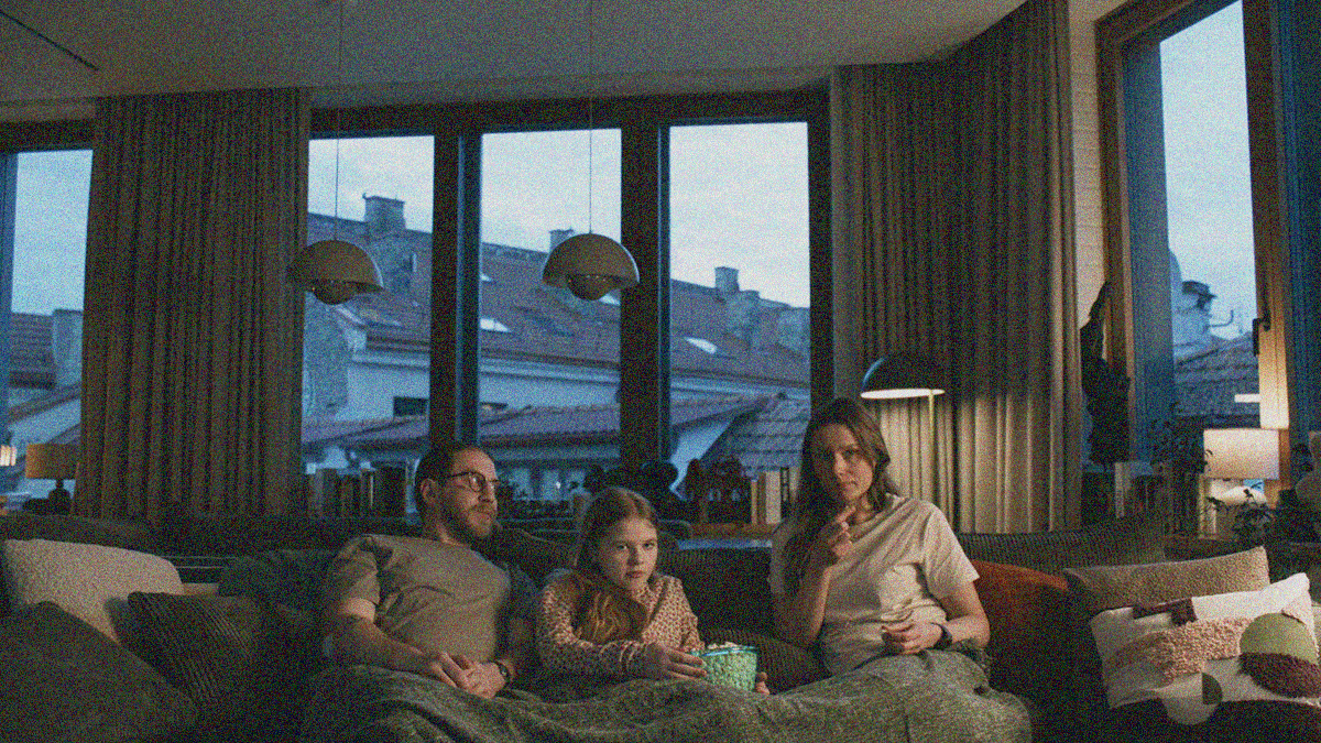 A father and mother bookend their daughter while sitting on the couch and eating popcorn.