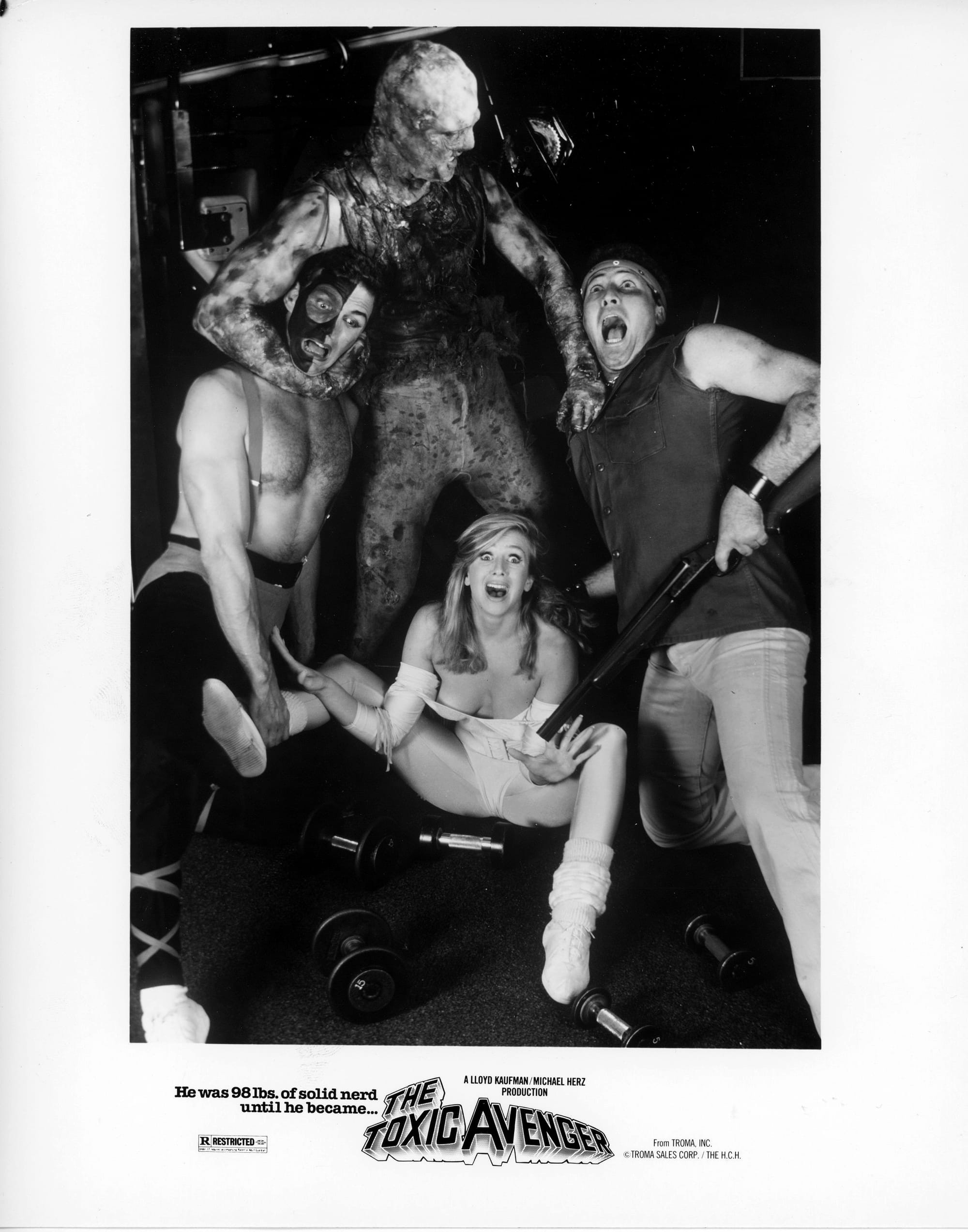 Publicity photo of the Toxic Avenger throttling two men in a weight room while a woman sits between them on the floor. All are screaming. Tagline: He was 98lbs. of solid nerd until he became ... THE TOXIC AVENGER A LLOYD KAUFMAN / MICHAEL HERZ PRODUCTION. From TROMA, INC © TROMA SALES CORP. / THE H.C.H