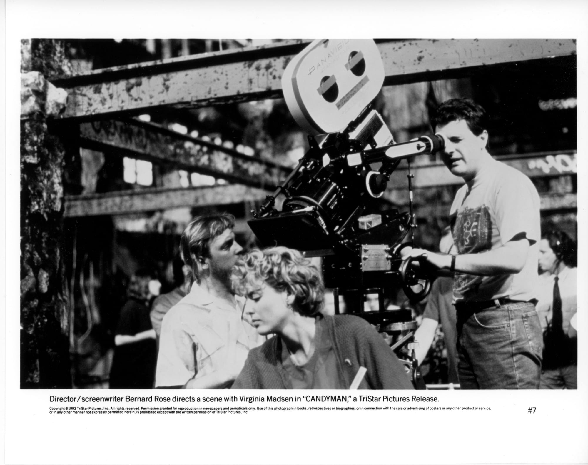 B&W Publicity Photo: A man in a t-shirt stands behind a movie camera. He looks through the viewfinder as it tilts downwards to capture a scene with a woman in the foreground.