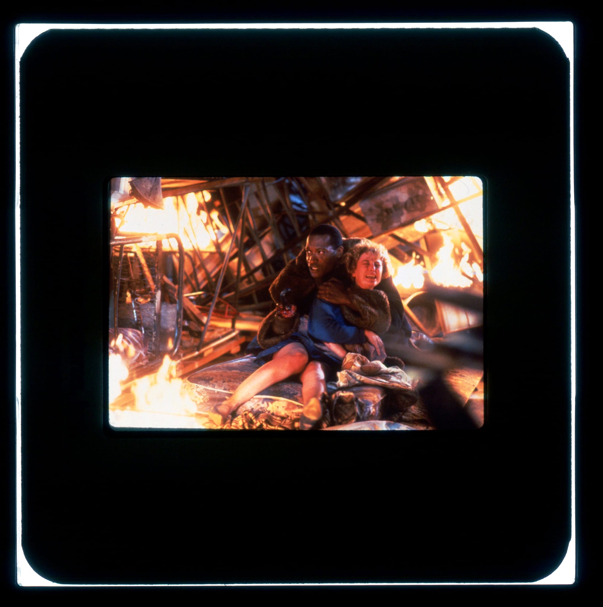 Color publicity slide: A man holds onto a screaming woman, both on the ground as their surroundings burn in fire.