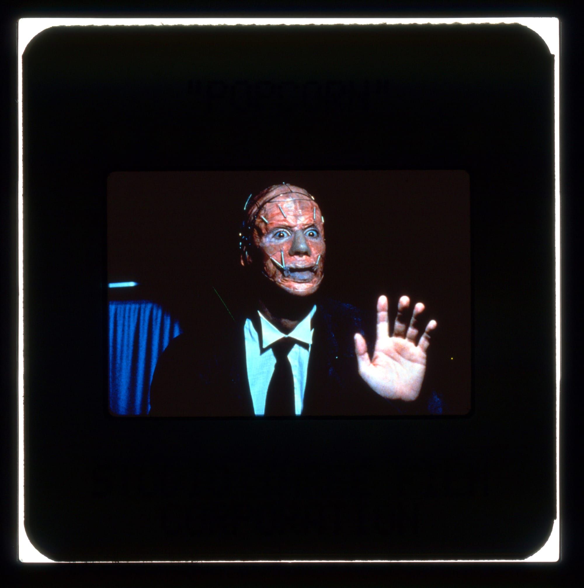 A man in a suit holds a hand up. His face is covered in a weird mask of bandages.