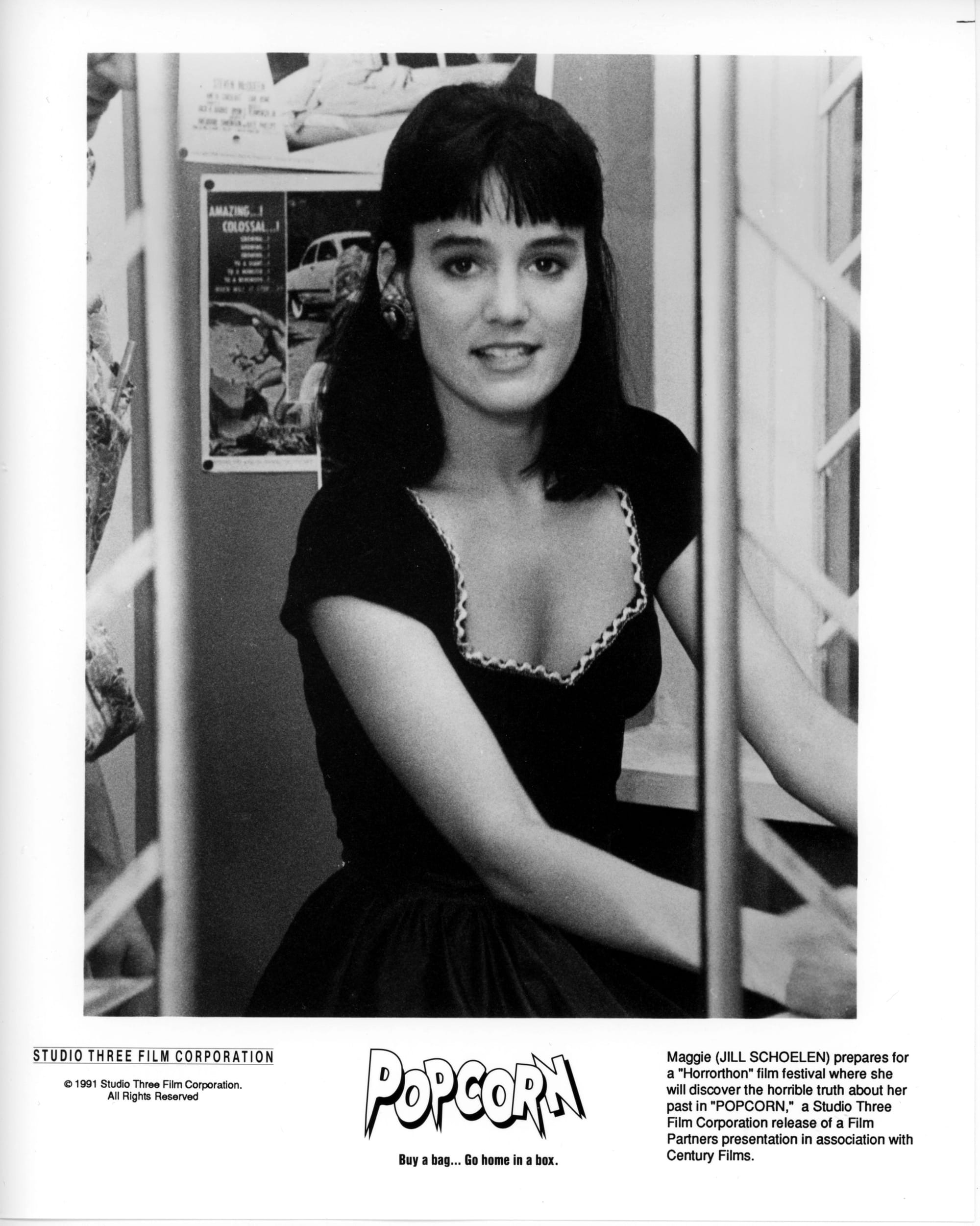A woman in a dress is seated in a room with photos tacked to the wall behind her.