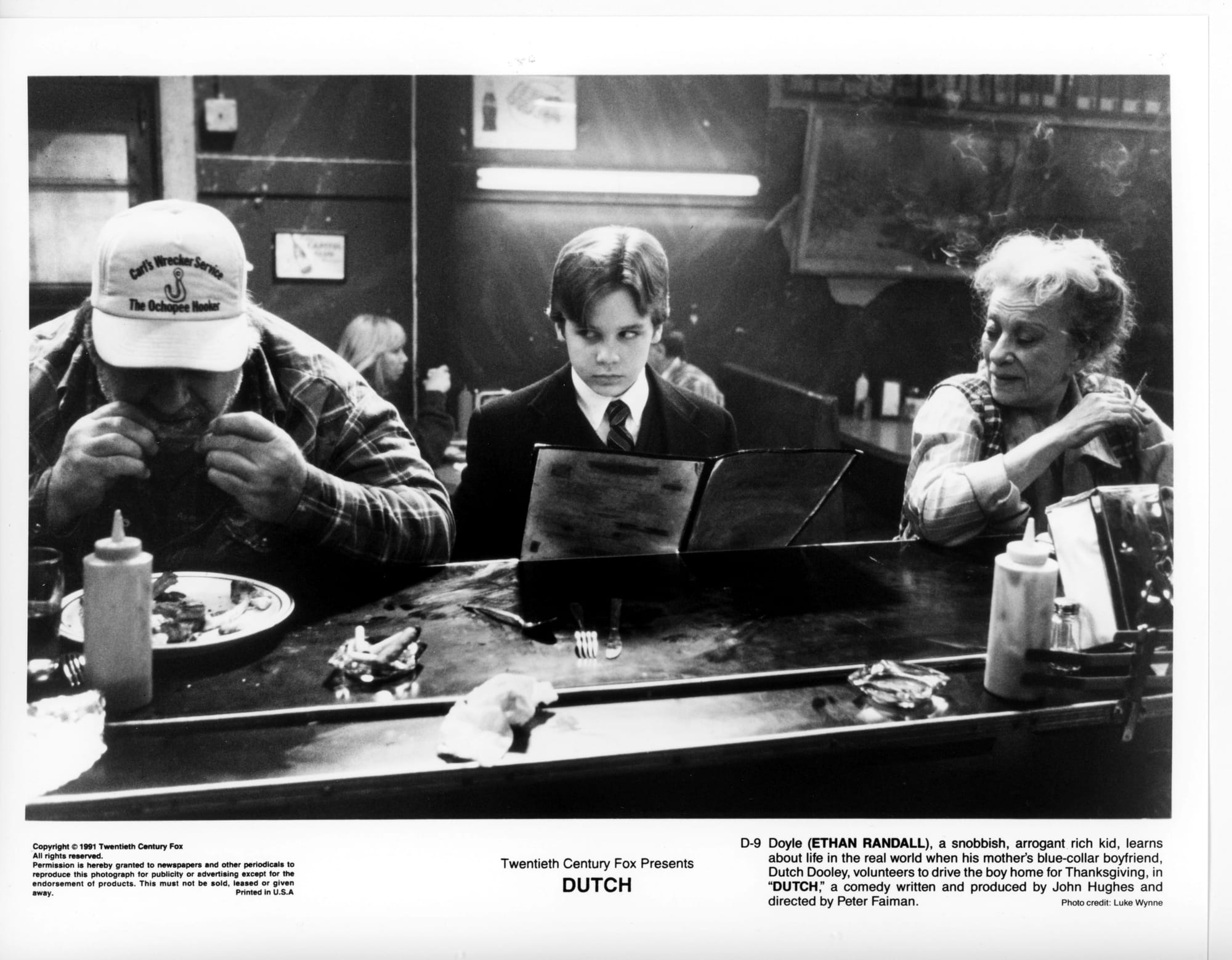 B&W publicity photo. A stern young boy sits at a dive bar counter with menu, side-eyeing a man to his right eating ribs.