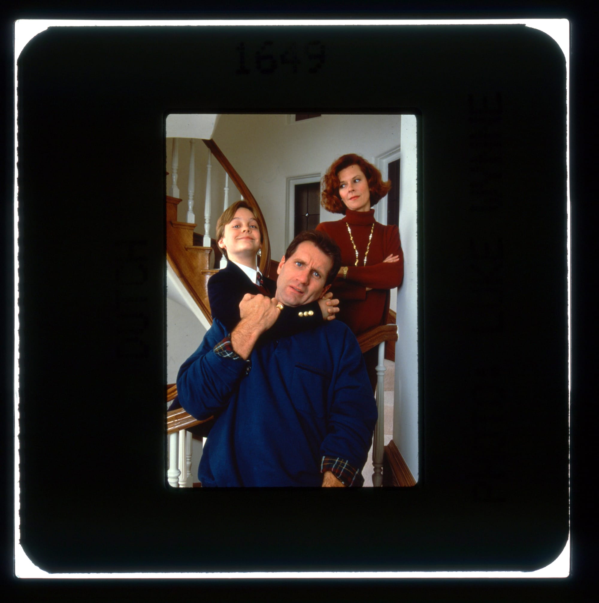 Color publicity slide of a family portrait of an annoyed man with a smirking boy hanging around his neck and judging mother standing behind with arms folded. 