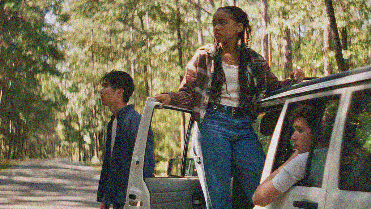 A shot of a white car on a forest road, cropped to the bottom right of the frame. One man stands at its front, a woman stands in the driver side door while balancing her arm on the roof, and another man sits in the backseat. All three are looking off-screen left.