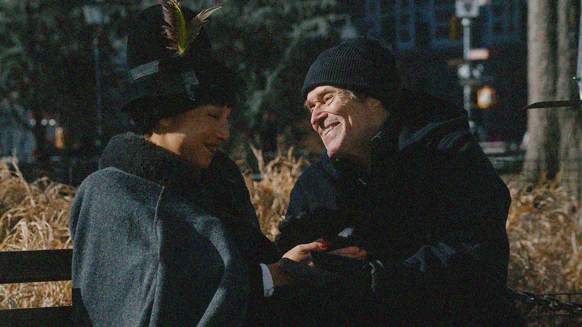 A woman in a big hat and an older man in winter cap sit on a bench, smiling and holding hands.