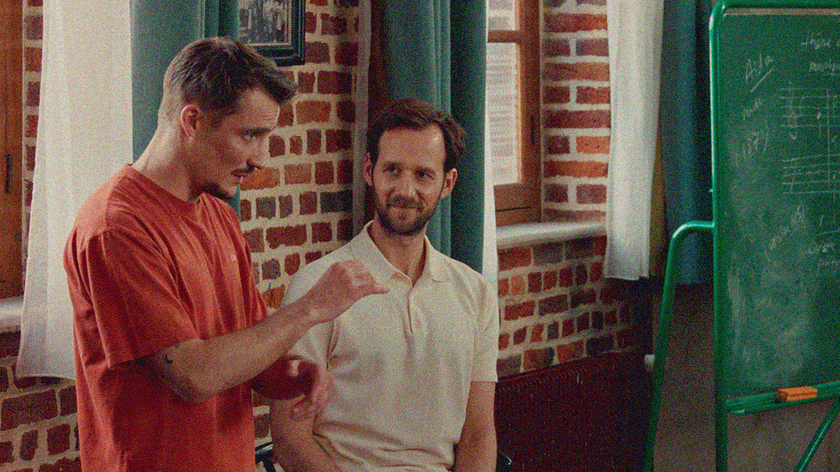A man in an orange tee at left conducts a band off-screen left while another man in a white polo looks on with a smile.